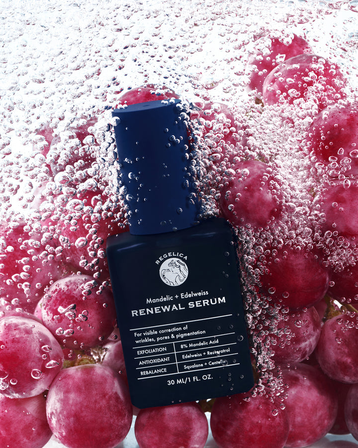 Regelica Mandelic + Edelweiss Renewal Serum in a dark navy glass pump bottle with grapes and water bubbles in the background